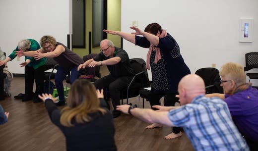 Candace and Daisy in a group session, demonstrating how to hover above a chair with arms by their ears and torse leaning forward. The students are mirroring them.