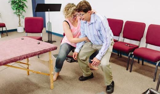 Candace helping a patient find a straight spinal position in a half squat position. She is standing to the right of the patient, with one hand holding the base of his neck, and the other hand supporting his torso