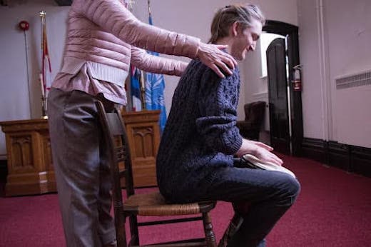 A patient is sitting on a chair and leaning forward at a 30 degree angle. The therapist is standing behind the chair with their hands on the patient's shoulders and assisting them.