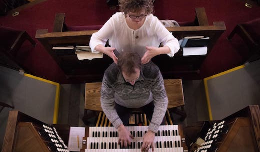 Aerial view of Candace standing behind a patient whie he plays a church organ. She is supporting the position of his head over his spine.